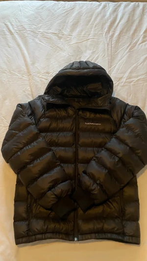 Svart pufferjacka från Peak Performance - Säljer en svart pufferjacka från Peak Performance med huva och dragkedja framtill. Jackan har en klassisk quiltad design och logga på bröstet. Perfekt för kalla vinterdagar och riktigt skön att bära. Materialet är glansigt och ger en modern look.