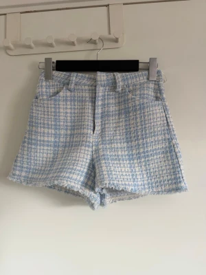 Blåvita tweedshorts med fransad kant - Säljer ett par blåvita tweedshorts med klassiskt rutmönster och fransad kant. Shortsen har fickor fram och bak samt hällor för skärp. Materialet är strukturerat och ger en trendig look. Perfekta för dig som vill ha något unikt och stilrent i garderoben.