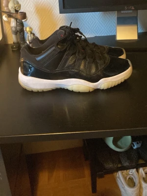 Svarta Air Jordan 11 Low sneakers - Säljer ett par svarta Air Jordan 11 Low sneakers med glansig patentläderdetalj, svart ovandel i läder och mesh samt vit mellansula och isiga, genomskinliga yttersulor. Klassisk siluett med coola detaljer och ikonisk design.