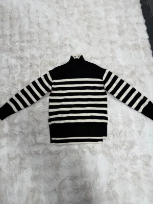 Svartvit randig stickad tröja - Snygg stickad tröja med svarta och vita horisontella ränder. Tröjan har hög krage, långa ärmar och ribbade muddar. Materialet ser mjukt och bekvämt ut, perfekt för kyligare dagar. Detalj med slits i sidan för extra stilpoäng.