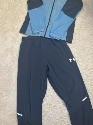 Under Armour träningsset mörk blå/ljusblå  - Snyggt träningsset från Under Armour med jacka och byxor i mörkblå och ljusblått. Jackan har hel dragkedja, logga på bröstet och elastiska muddar. Byxorna är svarta med logga på låret och reflexdetaljer vid benslut. Perfekt för löpning eller gym. Byxorna är i storlek xs och överdelen i storlek S . Men kan även passa i m beror på läng o vikt 
