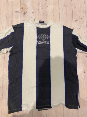 Randig t-shirt från Sweet Sktbs x Umbro - Säljer en randig t-shirt från Sweet Sktbs i samarbete med Umbro. T-shirten har breda vertikala ränder i vitt, svart och marinblått samt Umbro-logga broderad på bröstet. Klassisk passform och rund hals. Perfekt för dig som gillar streetwear.