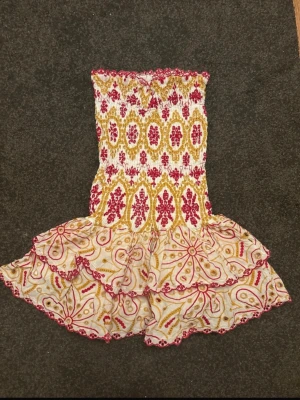 Mönstrad kortklänning med volang - Supersöt kortklänning i vitt med gult och rött mönster. Klänningen är axelbandslös och har smock upptill samt volangig kjol. Perfekt för varma dagar och sommaren. Materialet känns som bomull och lite av mönstret är broderat för extra detalj. Orginal pris 700kr