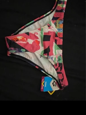 Färgglad bikiniunderdel från Arena - Säljer en färgglad bikiniunderdel från arena med grafiskt mönster i rosa, rött, grönt, blått och svart. Modellen har svarta knytband i sidorna för justerbar passform. Perfekt för stranden eller poolen!