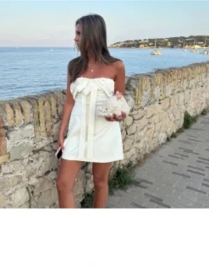 Vit axelbandslös kortklänning med rosett - Supersöt vit axelbandslös kortklänning med stor rosettdetalj framtill. Klänningen har en rak, något A-linjeformad siluett och är perfekt för sommarens alla tillfällen. Tillverkad i ett lätt tyg som ger en luftig känsla. Från Zara storlek M!