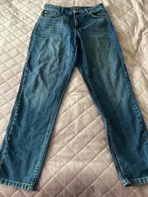 Blå raka jeans med klassisk passform - Ett par blå jeans med rak passform och klassisk femficksdesign. Jeansen har en lätt tvättad look med subtila slitningar framtill och normal midja. Tillverkade i denim, perfekta för en avslappnad stil. Dragkedjegylf och knapp i midjan.