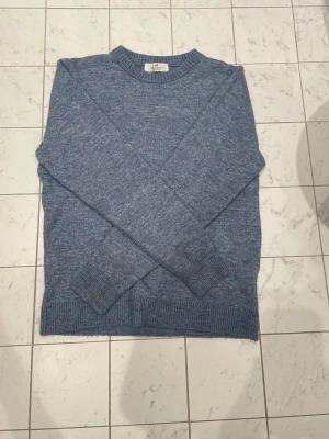 Blå stickad tröja från Arket - Mysig blå stickad tröja från Arket i mjukt material. Tröjan har rund halsringning, ribbade muddar vid ärmslut och nederkant samt en klassisk rak passform. Perfekt för kyliga dagar och enkel att matcha med olika outfits.