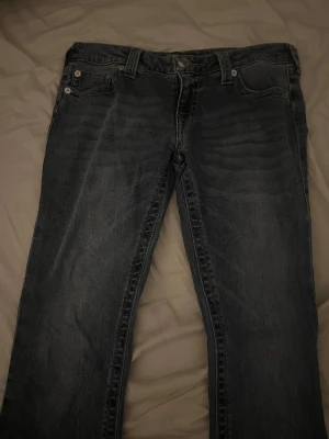 Mörkblå True Religion bootcut jeans - Säljer ett par mörkblå bootcut jeans med klassisk femficksdesign och snygga slitningar framtill. Jeansen har normal midja och är tillverkade i ett mjukt denimtyg. Perfekta för dig som gillar en avslappnad men trendig look.