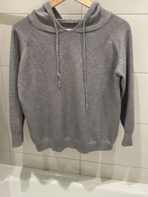 Grå stickad hoodie med dragsko - Mysig grå hoodie i stickat material med huva och dragsko. Långa ärmar och ribbade muddar vid ärmslut och nederkant. Perfekt för en avslappnad och stilren look.