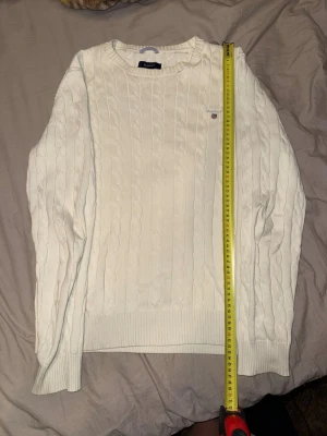 Vit kabelstickad tröja från GANT - Kabelstickad vit tröja från GANT med rund halsringning och broderad logotyp på bröstet. Tröjan har ribbade muddar vid ärmslut och nederkant, samt klassisk kabelstickad struktur som ger en tidlös look.