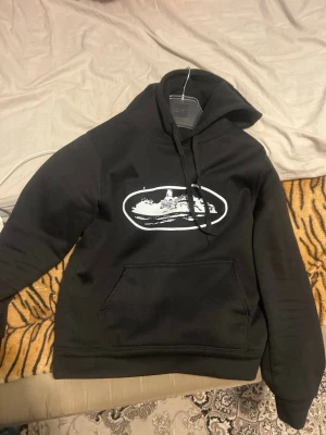 Svart hoodie med vit grafiskt tryck - Säljer en svart hoodie med ett vitt grafiskt tryck på bröstet som föreställer ett fartyg. Hoodien har klassisk känguruficka framtill och justerbar dragsko i huvan. Perfekt för dig som gillar streetwear och vill ha en unik look.