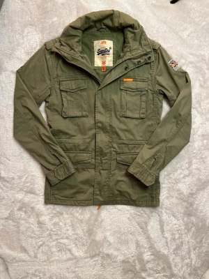 Superdry Field Jacket S - Tja! Säljer nu denna sjukt feta Field Jacket från Superdry. Den är i storlek S, riktigt najs passform. Perfekt nu till våren och sommaren. Dragkedjan är sönder, går säkert att laga, men det finns knappar så ingen fara. Hör av dig om du undrar något! 