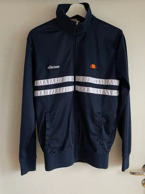 Marinblå track jacket från Ellesse - Snygg marinblå track jacket från Ellesse med vita dubbla ränder över bröstet och dragkedja hela vägen. Tröjan har hög krage, två fickor fram och broderad logga på bröstet. Perfekt för en sportig och retro look.