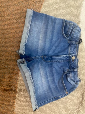 Blå jeansshorts med fransig kant - Säljer ett par blå jeansshorts med fransig kant och klassisk femficksmodell. Shortsen har knapp och dragkedja framtill samt bälteshällor. Perfekta för sommaren och har en avslappnad, trendig look.