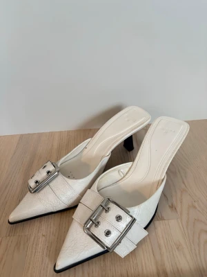 Vita pumps med stor silverdetalj - Snygga vita pumps med spetsig tå och bred rem med stor silverfärgad spänne. Skorna har öppen häl och smal klack, vilket ger en trendig och elegant look. Tillverkade i ett glansigt material som liknar läder.