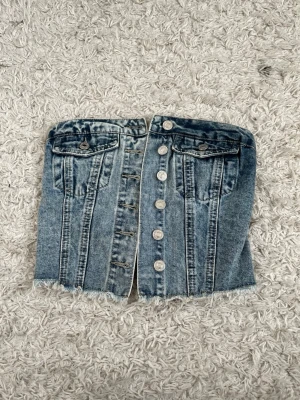 Blå denim korsett med knappar - Trendig blå denim korsett med framknäppning och rå kant nertill. Toppen har dekorativa fickor framtill och silverfärgade knappar. Perfekt för att skapa en edgy look till din outfit.