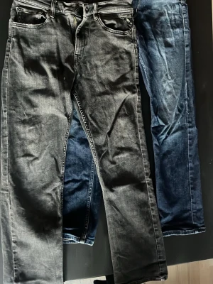 Svarta och blåa jeans från Replay - Två par jeans från Replay, ett par svarta och ett par blåa. Klassisk femficksmodell med dragkedja och knapp. Jeansen har raka ben och tydliga sömmar. Perfekta för dig som gillar en avslappnad och stilren look. Lite paj vid fickan som man ser på tredje bilden. 