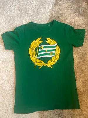 Grön Hammarby T-shirt - Snygg grön t-shirt från Hammarby IF med stort klubbmärke i gult, vitt och grönt på bröstet. Klassisk rund hals och korta ärmar. Perfekt för dig som vill visa ditt stöd för Bajen!