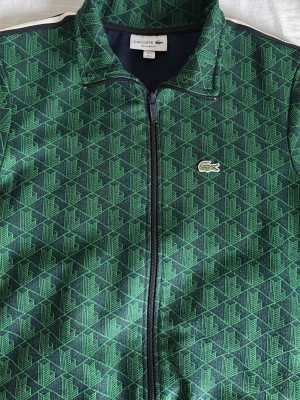 Grön mönstrad hoodie från Lacoste - Säljer en grön hoodie från Lacoste med hel dragkedja och ett grafiskt mönster i grönt och svart. Klassisk Lacoste-krokodil på bröstet och vita/gröna ränder på ärmen. Skön passform och sportig look, perfekt till streetwear.