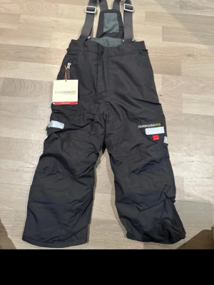 Svarta täckbyxor Didriksons med hängslen - Svarta täckbyxor från Didriksons med justerbara hängslen och reflexdetaljer. Byxorna har hög midja, dragkedja framtill och förstärkta partier på knän och benslut. Perfekta för kalla dagar och utomhusaktiviteter.