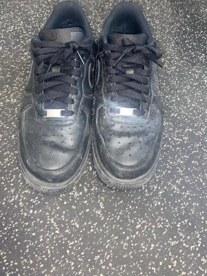 Svarta Nike Air Force 1 sneakers - Klassiska helsvarta Nike Air Force 1 sneakers i läder med svart sula, svarta skosnören och diskret Nike-logga på sidan. Modellen har perforeringar på tån för ventilation och metallbricka på snörningen. Perfekt för dig som gillar tidlös streetstil. Dom är dock lite sönder inuti men man ser inte de och jätte lite på utsidan 