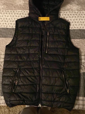 Svart parajumper väst - Snygg svart dunväst med huva och gul detalj vid halsen. Västen har tre dragkedjeförsedda fickor framtill och quiltad design. Perfekt för lager-på-lager och streetwear-stil. Materialet är glansigt och ger en modern look.