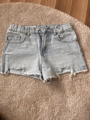 Ljusblå jeansshorts - Säljer ett par ljusblå jeansshorts från Zara med råa, fransiga kanter och slitna detaljer framtill och backtill. Klassisk femficksmodell med lite slitna detaljer på. Perfekta för sommaren och ger en avslappnad vibe.