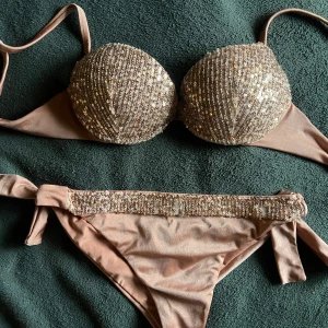 Glittrig beige bikini från Calzedonia - Supersnygg beige bikini från Calzedonia med glittriga paljettdetaljer på både överdel och nederdel. Överdelen har vadderade kupor och knyt i ryggen, medan underdelen har knyt i sidorna och matchande paljetter framtill. Perfekt för sommarens strandhäng! Som man ser på bilden har litr av paljetterna lossnat på bikiniunderdelen!! Ungefär B-C kupa på toppen och Xs-S underdel ❤️