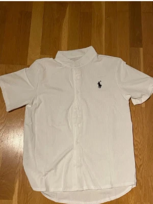 Vit kortärmad skjorta Polo Ralph Lauren - Säljer en vit kortärmad skjorta Polo Ralph Lauren med klassisk krage och knappar framtill. Skjortan har diskret randigt mönster och den ikoniska loggan broderad på bröstet. Perfekt till sommarens alla tillfällen!