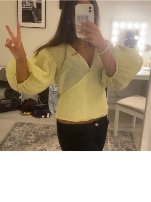 Gul omlottblus med puffärmar - Trendig gul omlottblus med stora puffärmar och v-ringning. Blusen har en lätt strukturerad yta och sitter tajt i midjan för en snygg siluett. 