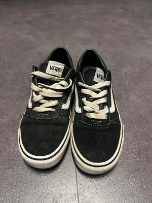 Svarta Vans Old Skool sneakers - Klassiska Vans Old Skool sneakers i svart mocka och canvas med vita detaljer och vit sula. Skorna har den ikoniska sidostripen och vita snören. Perfekta för en avslappnad streetwear-look.