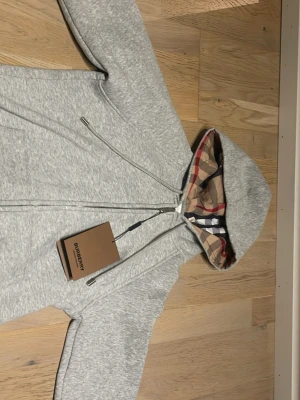 Grå zip hoodie från Burberry -  Snygg gråmelerad hoodie från Burberry med dragkedja och klassiskt rutigt foder i huvan. Hoodien har justerbara snören vid huvan och är tillverkad i mjukt bomullsmaterial. Perfekt för en avslappnad och trendig look.OPS är i liten storlek så passar som S/XS