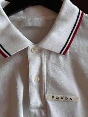 Vit pikétröja från Prada - Säljer en stilren vit pikétröja från Prada med klassisk krage och knäppning framtill. Kragen har snygga röda och svarta ränder som ger en sportig touch. Materialet är pikétyg i bomull och logotypen sitter diskret på bröstet. Perfekt för dig som gillar exklusiva märken.