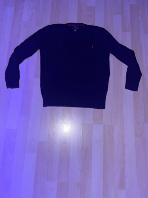 Mörkblå kabelstickad tröja Polo Ralph Lauren - Snygg mörkblå kabelstickad tröja från Polo Ralph Lauren med den klassiska röda loggan broderad på bröstet. Tröjan har rund halsringning och ribbade muddar vid ärmslut och nederkant. Perfekt för lager på lager och stilren look.
