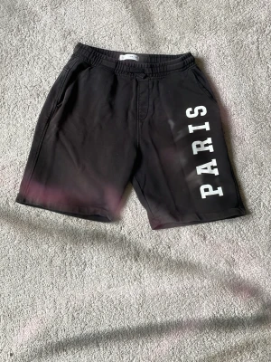 Svarta shorts med PARIS-tryck - Säljer ett par svarta shorts med resår i midjan och fickor på sidorna. På ena benet står det 'PARIS' i stora vita bokstäver. Sköna och avslappnade, perfekta för sommaren eller chill dagar.61kr frakt betalas av köpare.