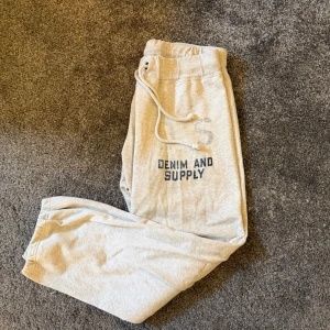 Grå mjukisbyxor Denim & Supply - Snygga ljusgrå mjukisbyxor från Denim & Supply med tryckt logga på ena benet. Byxorna har dragsko i midjan och resår vid bensluten för en skön och avslappnad passform. Tillverkade i mjukt bomullsmaterial som känns riktigt bekvämt. Inga defekter finns på plagget.
