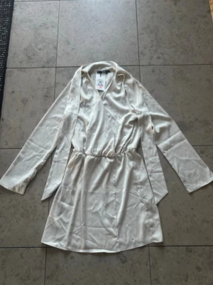 Ljus omlottklänning från BikBok - Ljus omlottklänning från BikBok i sidenliknande material. Klänningen har långa ärmar, knytband i midjan och en mjuk, glansig finish. Perfekt för dig som gillar en stilren och elegant look.