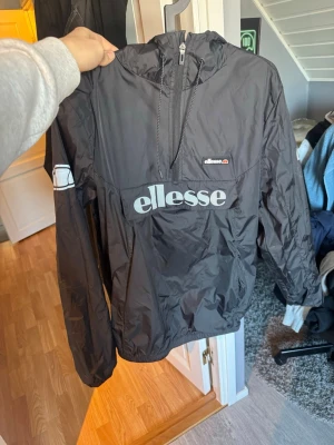 Svart vindjacka från Ellesse - Svart vindjacka från Ellesse med stor logga framtill och halv dragkedja. Jackan har huva, lång ärm och en stor ficka på magen. Tillverkad i lätt och glansigt material som skyddar mot vind. Perfekt för en sportig och avslappnad stil.