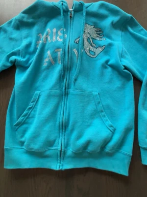 Rhinestone hoodie (ljusblå) - Turkos rhinestone hoodie dkorerad med glittrande stenar som bildar text och logga. Mjuk bomullsblandning och huva med dragsko. Perfekt för en chill och trendig look.