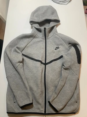 Grå Nike hoodie med dragkedja - Snygg grå hoodie från Nike tech fleece med hel dragkedja framtill och svarta detaljer. Hoodien har en huva, svarta linjer över bröstet och en ficka med dragkedja på ena ärmen. Perfekt för en sportig och avslappnad stil.