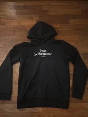 Svart hoodie Peak Performance - Snygg svart hoodie från Peak Performance med vit logga på bröstet. Klassisk modell med huva och känguruficka framtill. Tillverkad i mjukt material som passar perfekt till chill eller streetwear. Designad i Sverige.
