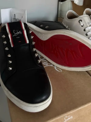 Christian Louboutins - ⚠️Byten mot gråa/mörkblåa junior spike är intressant⚠️, Svarta sneakers från Christian Louboutin i skinn med ikonisk röd sula och silverfärgade nitar längs sidorna. Tungan har en svart, röd och vit detalj med logga. Snygg och edgy design som sticker ut. Kvitto, dustbag och box följer med. Kan mötas i Stockholms området