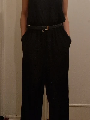 Svart ärmlös jumpsuit med bälte - Snygg svart jumpsuit med smala axelband och rak halsringning. Den har en clean och stilren look. Bältet ingår ej. 