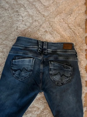 Blå Pepe jeans med tvättad look - Säljer ett par blå jeans från Peppe  med tvättad effekt och klassiska bakfickor med vågig söm. Jeansen har normal passform, raka ben och dubbla knappar i midjan. Perfekta för dig som gillar en avslappnad men snygg stil.