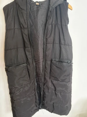Svart lång dunväst med huva - Snygg svart lång dunväst med huva och två stora fickor med dragkedja framtill. Västen har quiltad design och stängs med dragkedja. Perfekt för lager-på-lager och streetstyle. Dragkedjan är sönder.