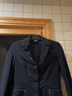 Mörkblå kavaj från Horse life  - Snygg mörkblå kavaj med klassisk krage, fyra knappar framtill och dragkedjefickor. Märke på ärmen. 