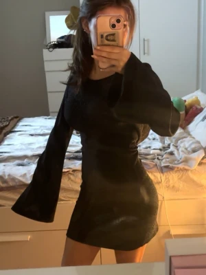 Svart kort klänning med vida ärmar - Snygg svart kort klänning med figurnära passform och vida, långa ärmar. Klänningen har en enkel design med rund halsringning och lätt glansigt material. Perfekt för dig som vill ha en stilren men ändå trendig look. Osäker på om jag vill sälja