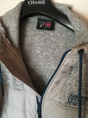 Grå jacka från Scandinavian Explorer - Mysig grå jacka från Scandinavian Explorer med huva och dragkedja. Jackan har blå detaljer, broderad logga på bröstet och patch på ärmen. Perfekt för kyliga dagar och har en sportig, avslappnad look.