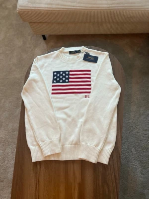 Vit stickad tröja Polo Ralph Lauren med USA flagga - Vit stickad tröja från Polo Ralph Lauren med broderad amerikansk flagga framtill.  • Storlek: L • Passform: Regular fit • Skick: Oanvänd – nyskick, med originaltag kvar • Färg: Vit • Detaljer: Rund halsringning, ribbade muddar och nederkant, broderad flagga samt diskret “RL” framtill  Tröjan är helt oanvänd och i perfekt skick. Klassisk Polo Ralph Lauren-design som funkar lika bra till vardag som mer uppklätt.  Nypris: ca 2 000–2 300 kr