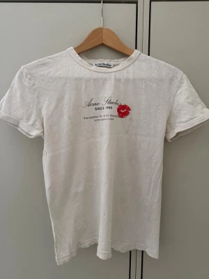 Vit Acne Studios t-shirt med tryck - Vit t-shirt från Acne Studios med svart texttryck och en röd kyssmunsdetalj på bröstet. Klassisk rund halsringning och korta ärmar. Tillverkad i mjuk bomull för en skön och avslappnad känsla. Perfekt till jeans eller kjol. Fläck på framsida, fotograferat andra fotot. 